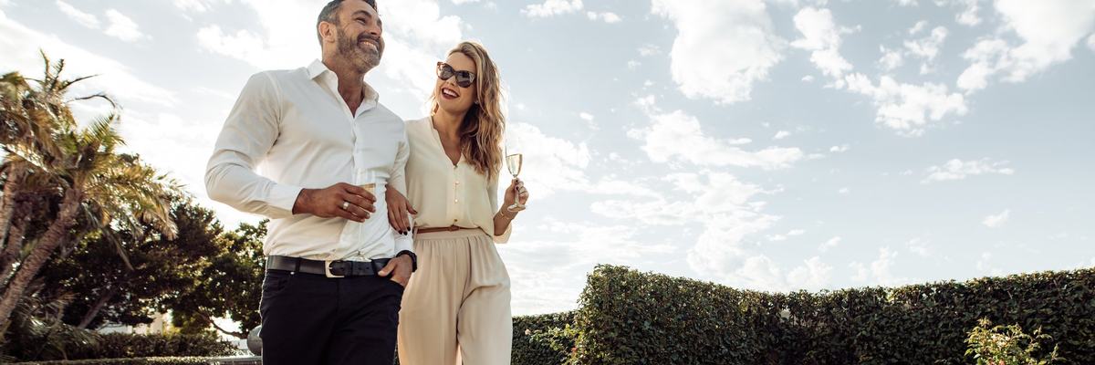 Well-dressed couple walking through greenery drinking champagne.