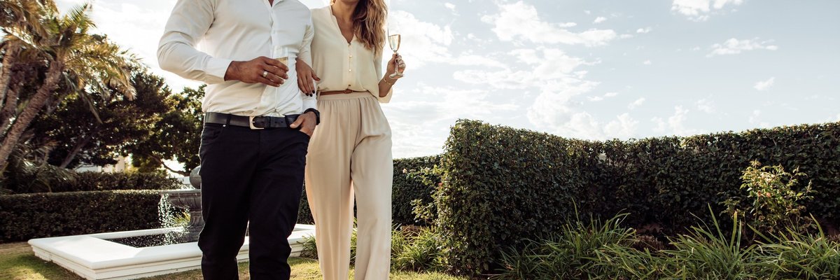 Well-dressed couple walking through greenery drinking champagne.