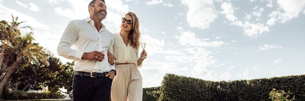 Well-dressed couple walking through greenery drinking champagne.