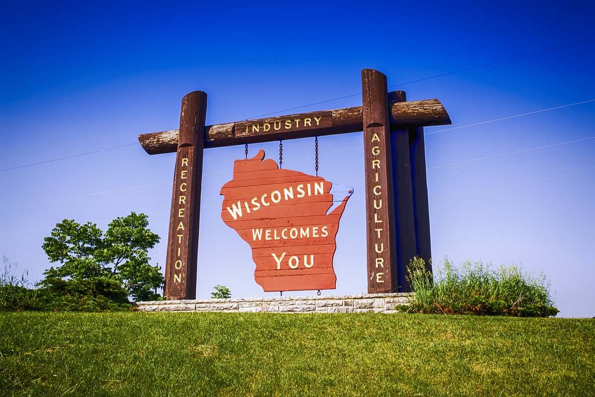 A large road-side sign surrounded by green grass and a blue sky welcoming visitors to Wisconsin.