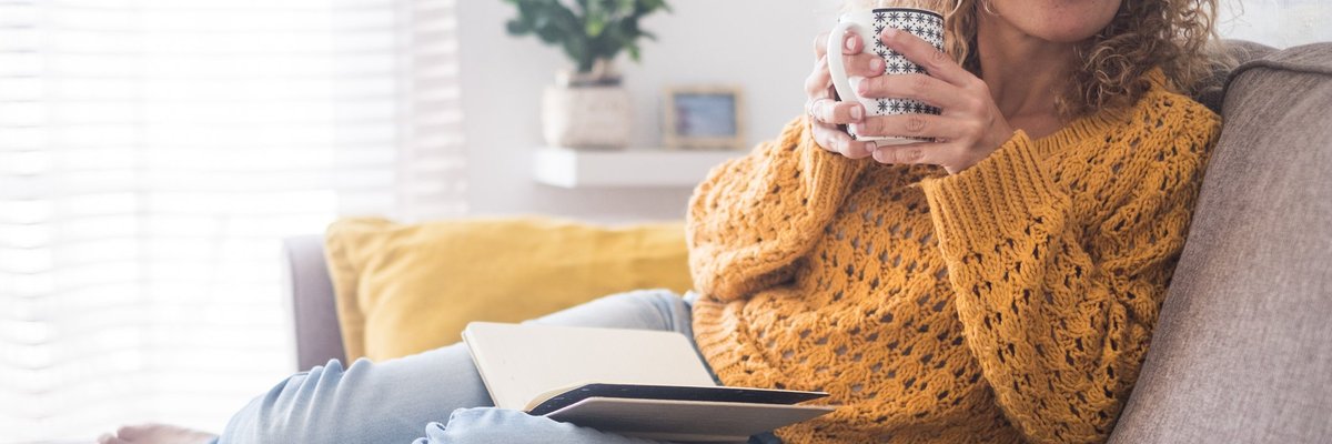 A woman drinking coffee and reading a book on a couch.