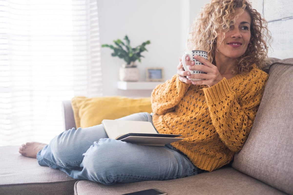 A woman sitting comfortably on her couch while drinking coffee and reading a book.