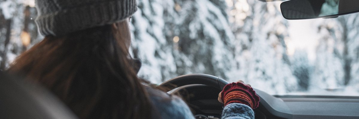 Woman in cold weather accesories driving a vehicle through snowy landscape.