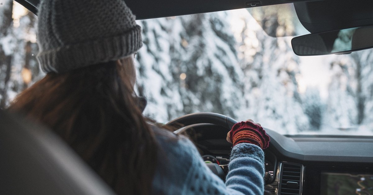 Woman in cold weather accesories driving a vehicle through snowy landscape.