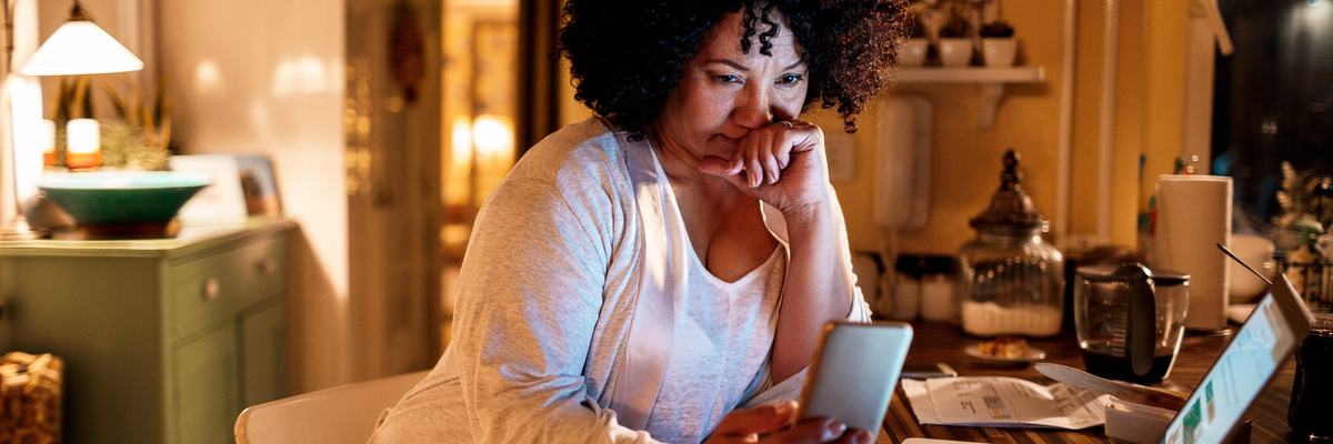 A woman in her kitchen sits at the island, using her phone and laptop.