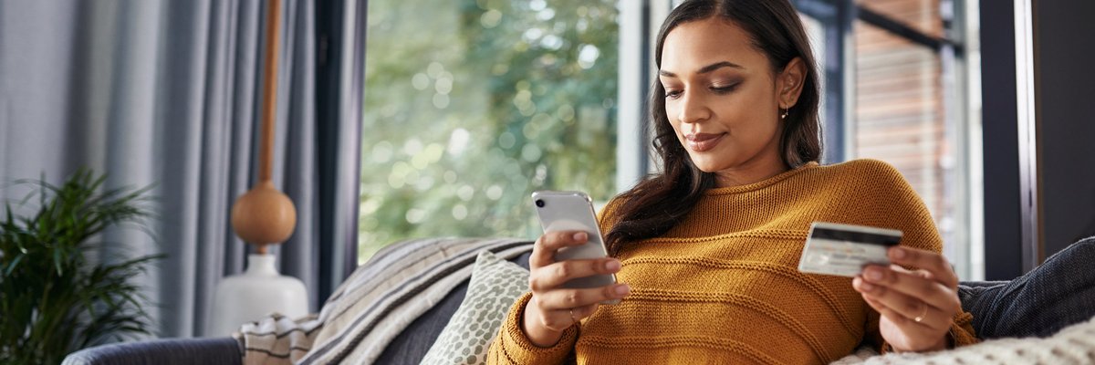 A woman sitting comfortably on her couch looking at her phone and holding a credit card.