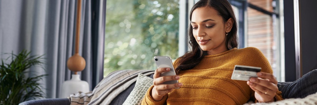 A woman sitting comfortably on her couch looking at her phone and holding a credit card.