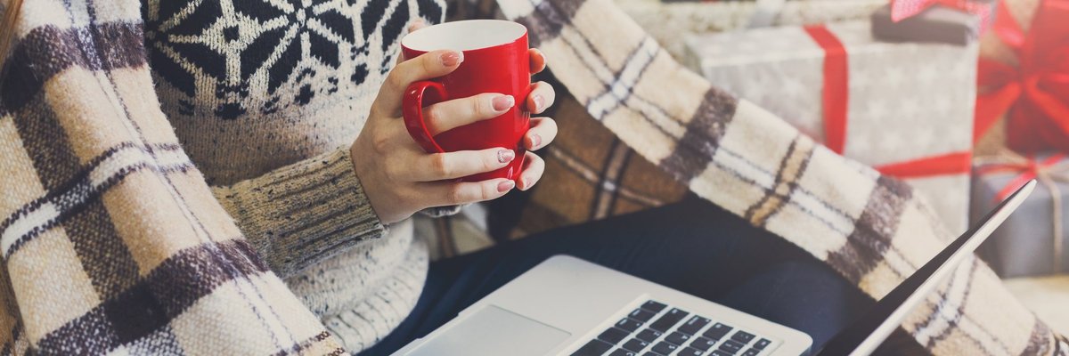 A woman holding a cup of coffee and a laptop with a blanket and presents around her.