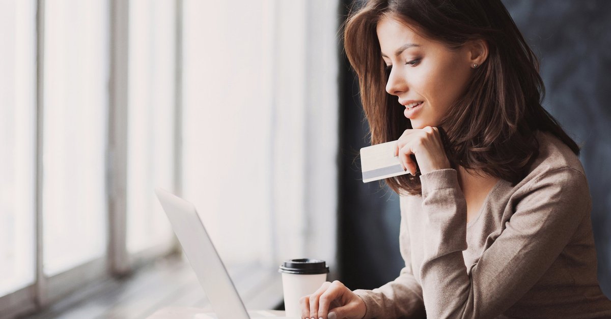A woman sitting in front of a window while typing on a laptop with one hand and holding a credit card with the other.