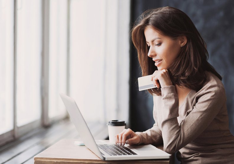 A woman sitting in front of a window while typing on a laptop with one hand and holding a credit card with the other.