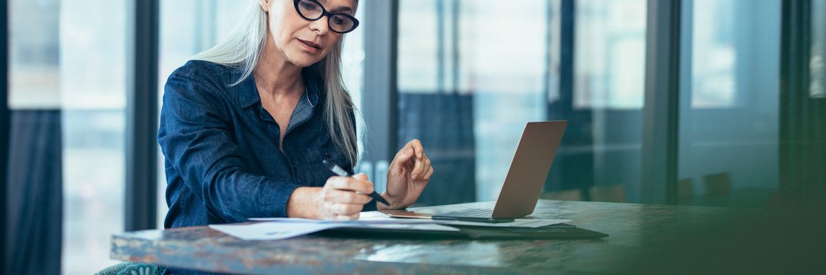 A woman reading paperwork at a table in front of large windows.