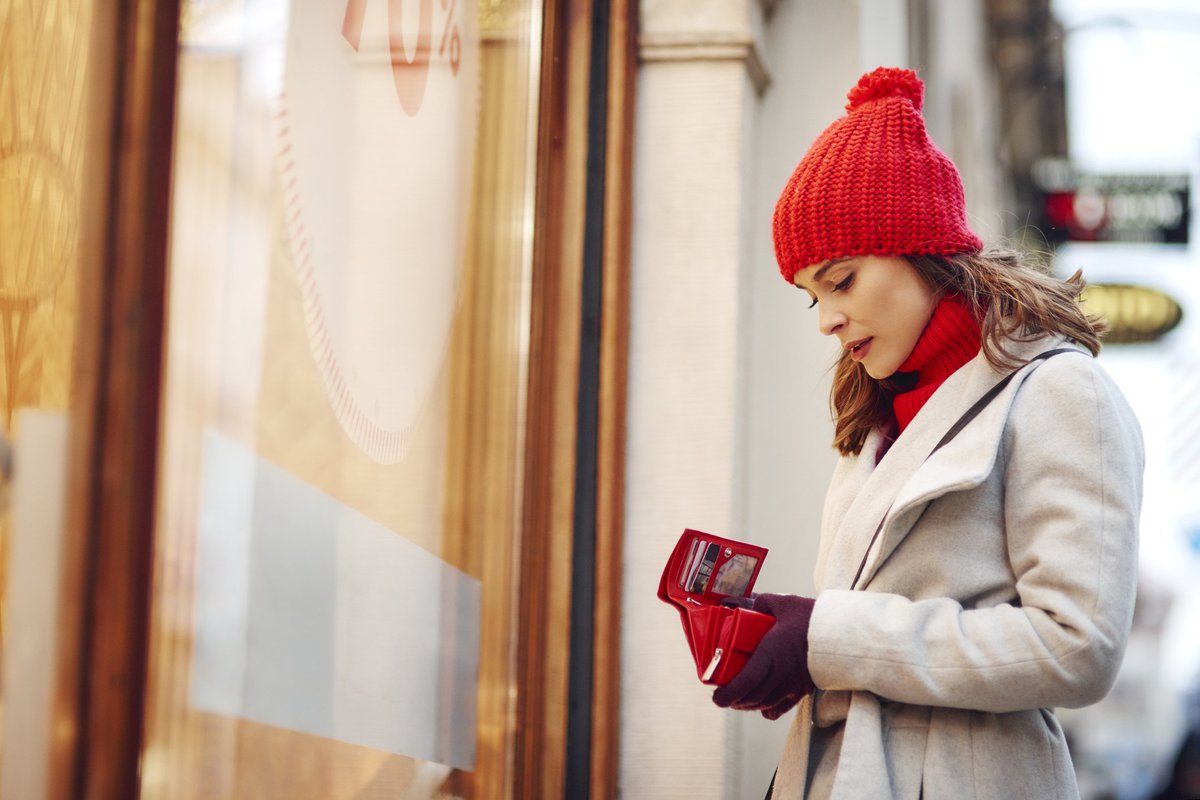 A woman in a winter hat looking at her empty wallet outside of a store.