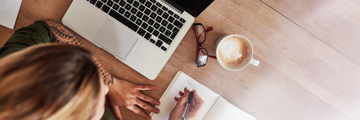 A woman writing out a to-do list at a desk next to a laptop and cup of coffee.