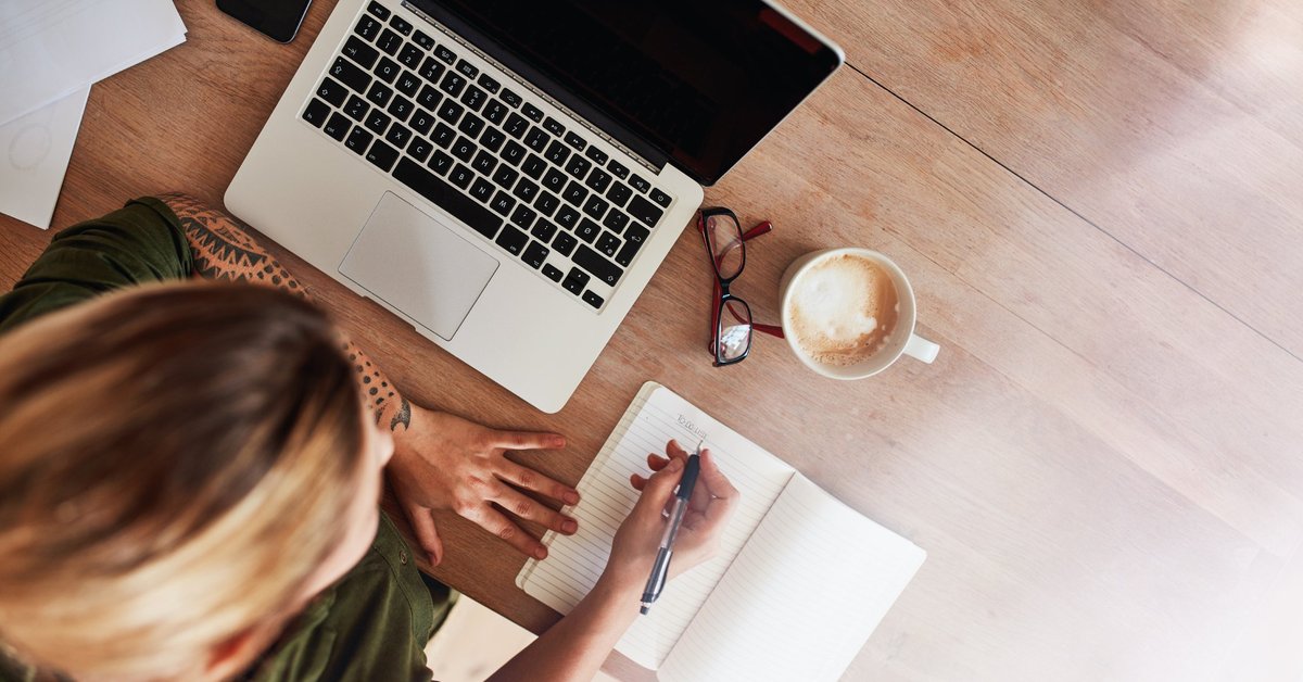 A woman writing out a to-do list at a desk next to a laptop and cup of coffee.