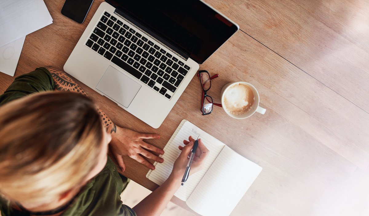 A woman writing out a to-do list at a desk next to a laptop and cup of coffee.