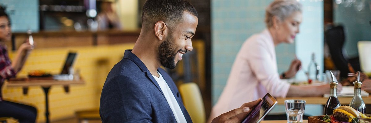 A young businessman sits in a coffee shop, reading on a tablet.