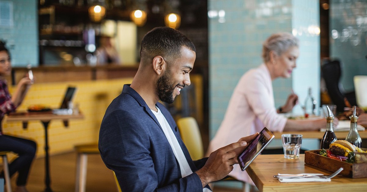 A young businessman sits in a coffee shop, reading on a tablet.