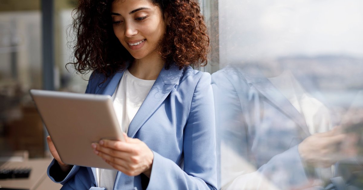 A young businesswoman leans against a window while looking at her tablet.