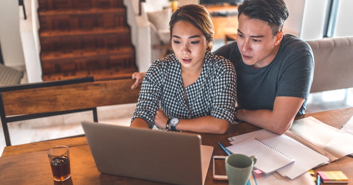 A serious young couple sitting at their dining table reading paperwork on their laptop.