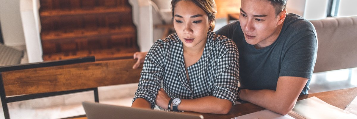 A serious young couple sitting at their dining table reading paperwork on their laptop.