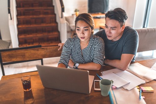 A young couple sitting at their dining table with notebooks and reviewing their finances on a laptop.