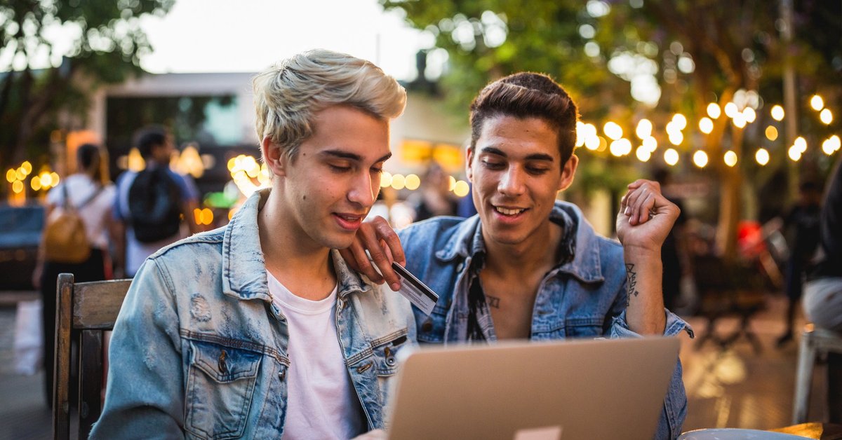 A young couple sit outside, looking at a computer. One is holding a credit card.