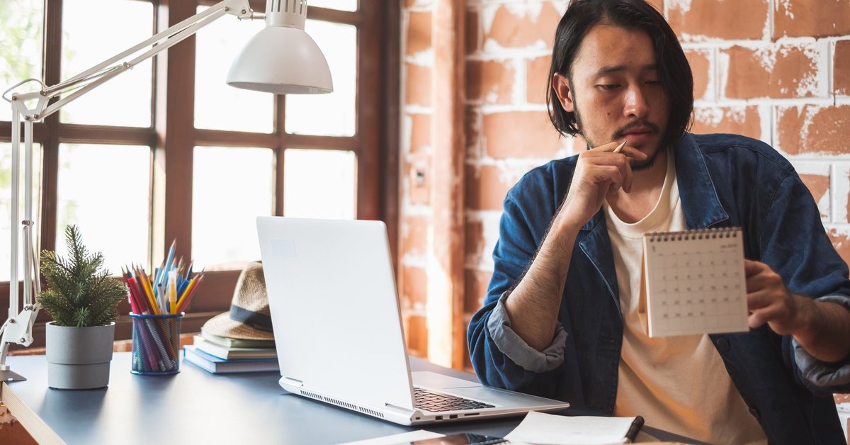 Young man pausing from using his laptop to look at a calendar.