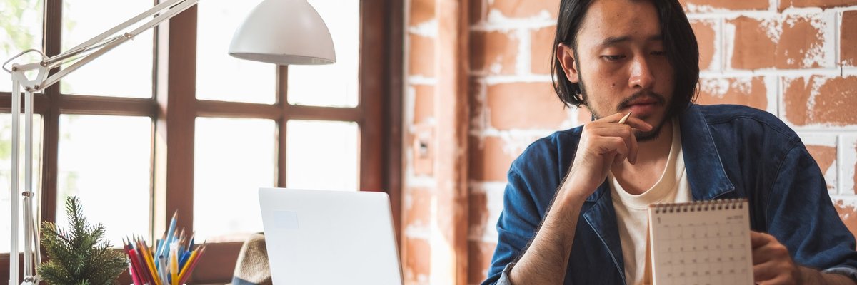 Young man pausing from using his laptop to look at a calendar.
