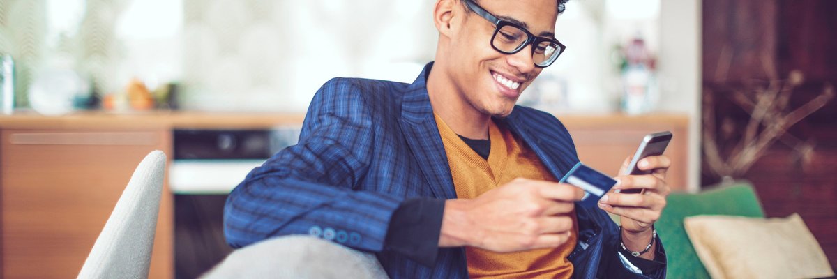 A young man, sitting on a sofa, smiles as he holds a credit card in one hand and looks at his cellphone in another.