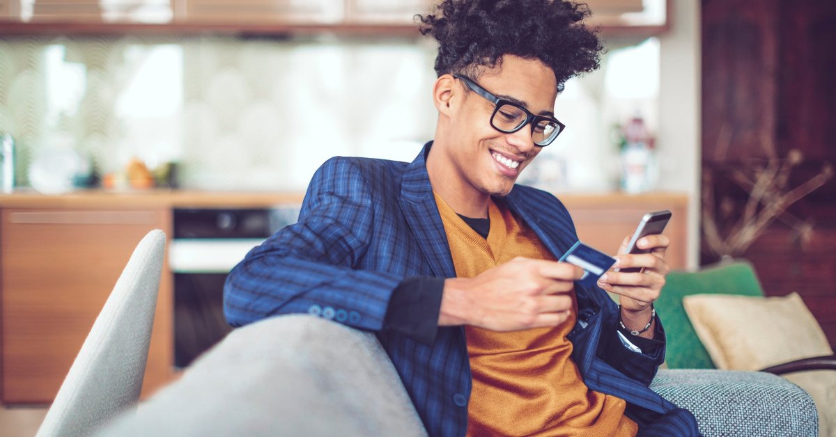A young man, sitting on a sofa, smiles as he holds a credit card in one hand and looks at his cellphone in another.