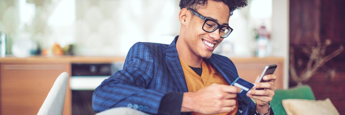 A young man, sitting on a sofa, smiles as he holds a credit card in one hand and looks at his cellphone in another.