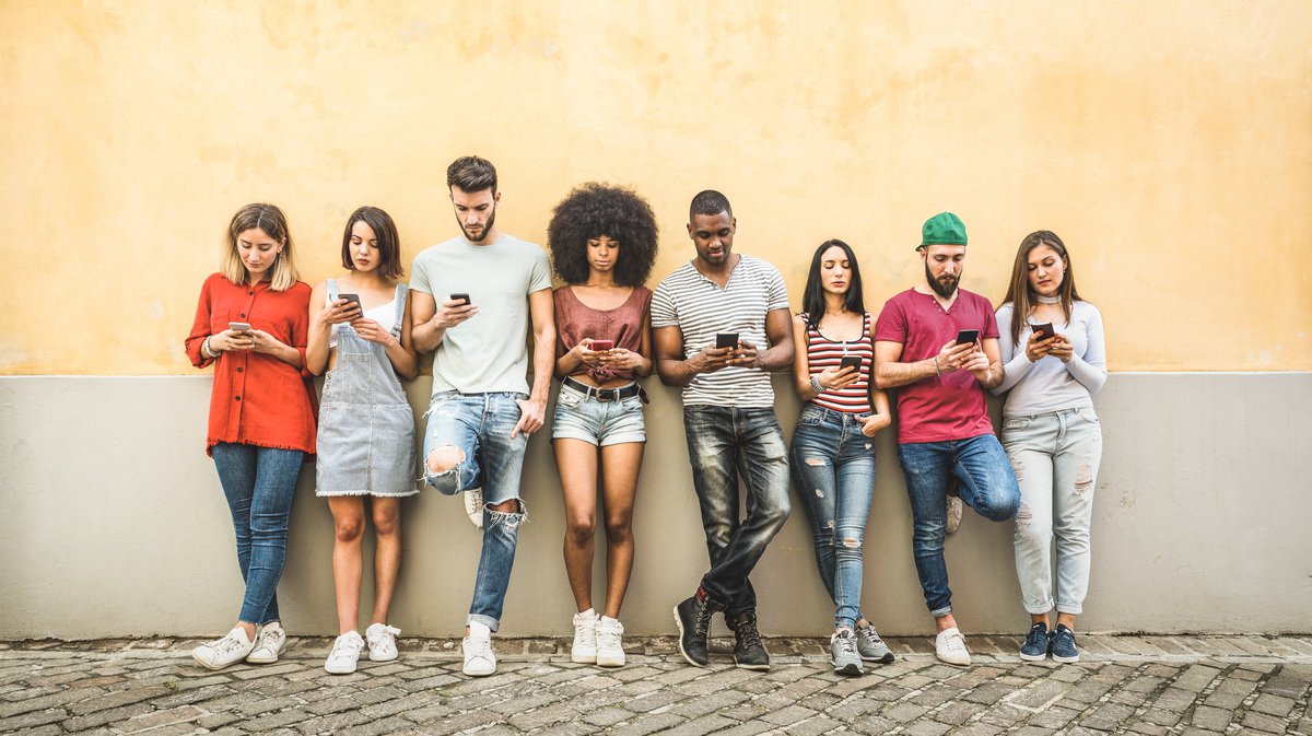 A group of young people leaning against a wall and all looking at their phones.