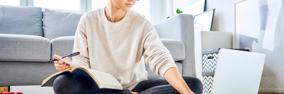 A young woman sitting on the floor of her living room with a notebook, calculator, and laptop.