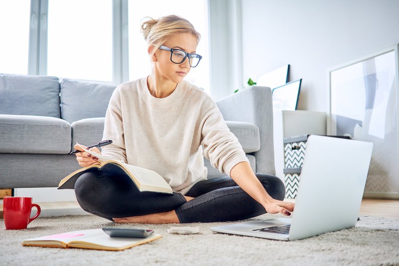 A young woman sitting on the floor of her living room with a notebook, calculator, and laptop.