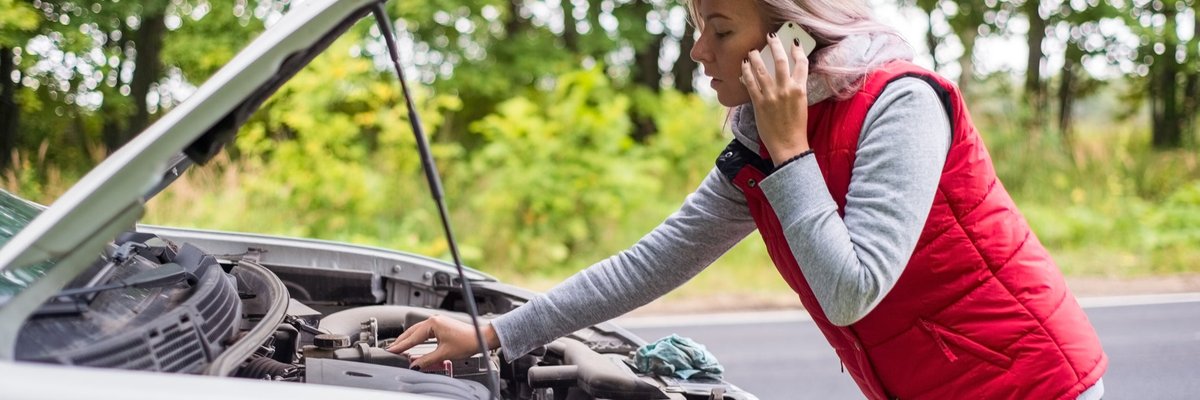 A young woman calls for road service while looking under the hood of her car.