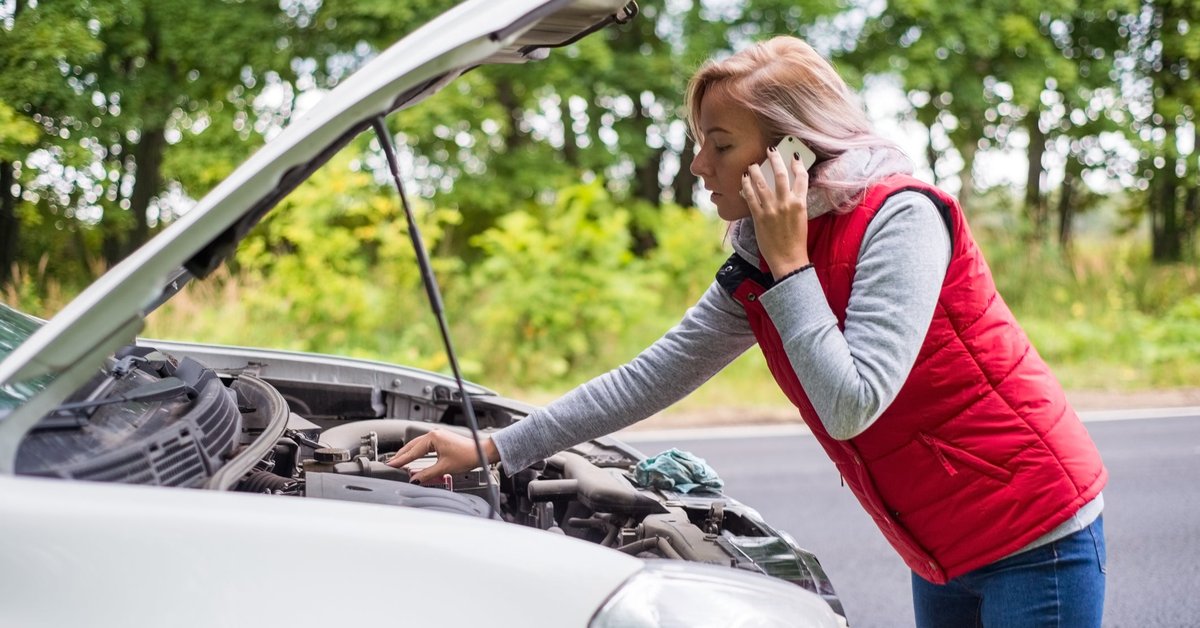 A young woman calls for road service while looking under the hood of her car.