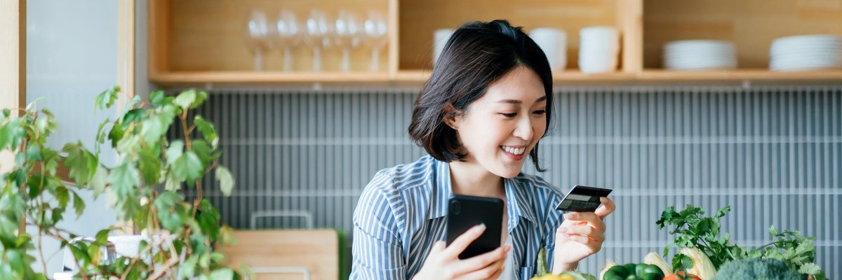 A young woman stands in her kitchen, holding her credit card and phone.