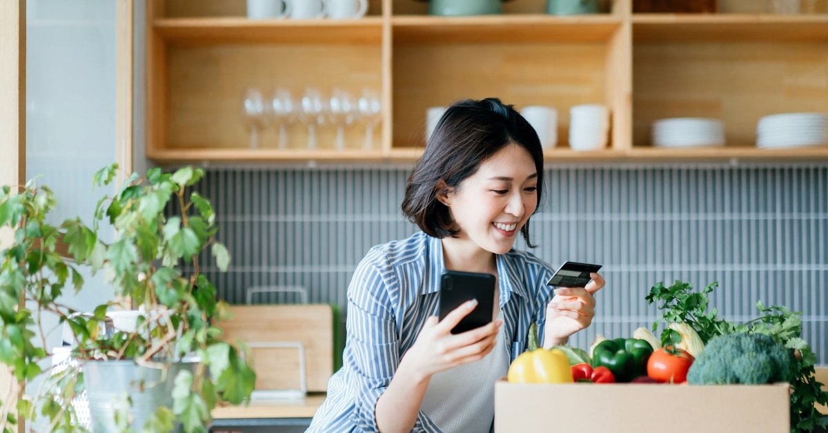 A young woman stands in her kitchen, holding her credit card and phone.