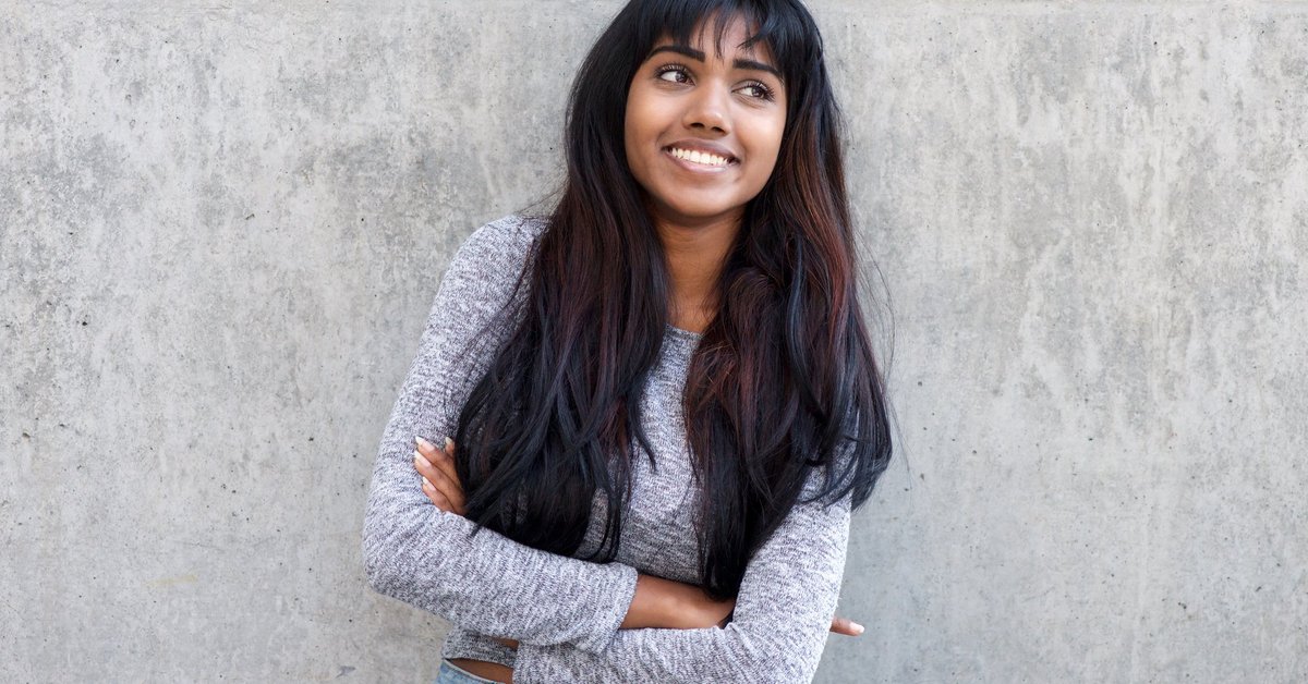 A smiling young woman standing against a wall with her arms crossed.