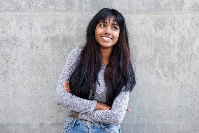 A smiling young woman standing against a wall with her arms crossed.