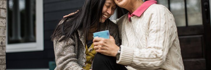 An adult woman and her senior mom sitting on their front porch steps while smiling and hugging.