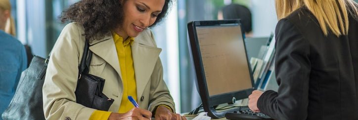 A person standing across from a teller at a bank counter and writing a check.