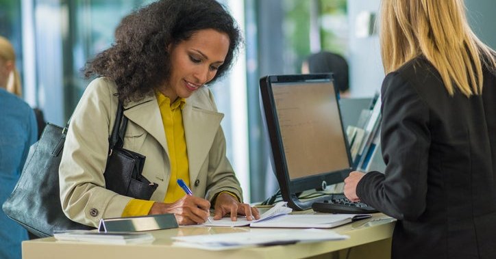 A person standing across from a teller at a bank counter and writing a check.