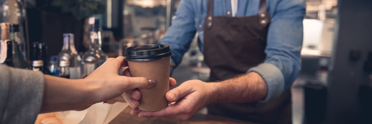 A barista handing a takeout coffee cup to a customer in a coffeeshop.
