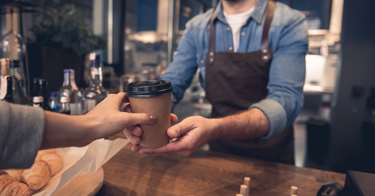 A barista handing a takeout coffee cup to a customer in a coffeeshop.