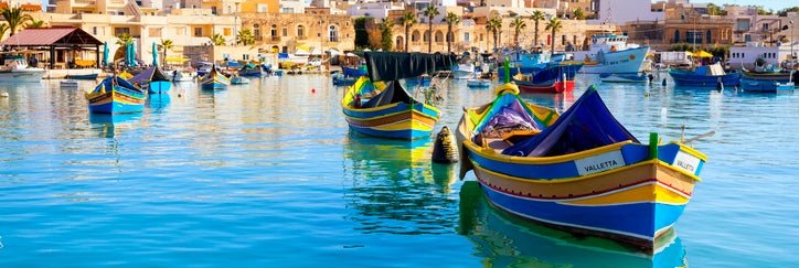 Colorful boats in a harbor with bright blue water with the Malta skyline in the background.
