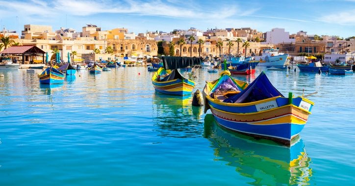 Colorful boats in a harbor with bright blue water with the Malta skyline in the background.