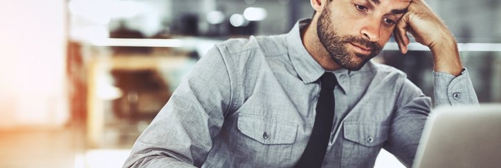 A man looking bored with his head resting on his hand while working on his computer in an office.