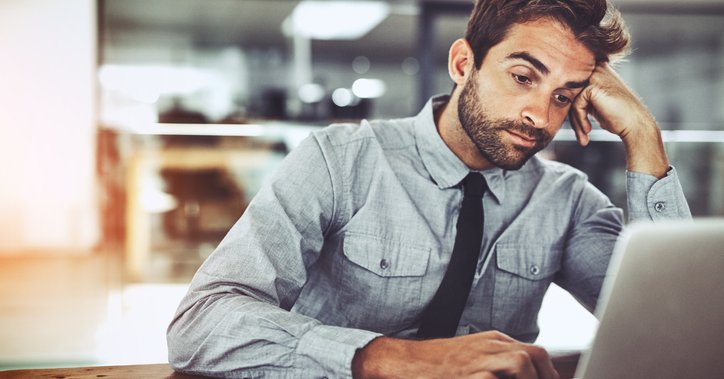 A man looking bored with his head resting on his hand while working on his computer in an office.