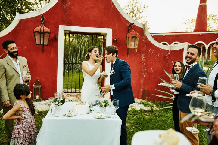 A bride feeding cake to a her groom at an outdoor wedding surrounded by guests.